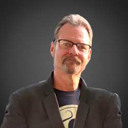 Headshot of Charles Minskoff wearing glasses, a blazer jacket and a tshirt and smiling